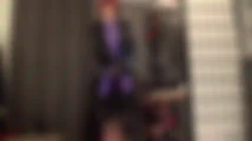 Indosso un cappotto in pvc pesante e lucido sopra una tuta viola in leohex con strapon stryker e stivali.