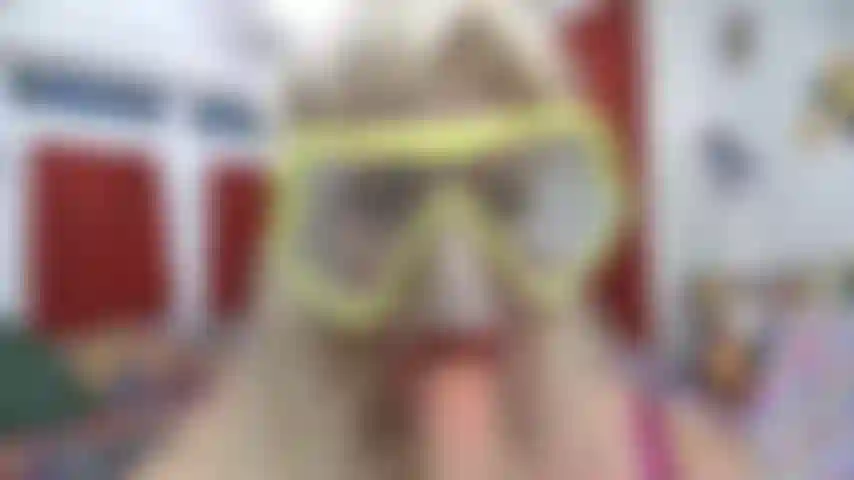 Con il viso in prospettiva e una maschera da sub appannata con un rossetto rosso che succhia un giocattolo bianco.